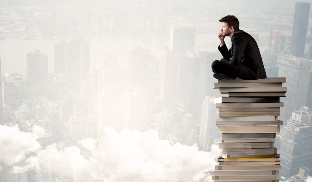 A serious businessman in elegant suit sitting on a pile of giant books in front of a grey city scape with clouds, fog